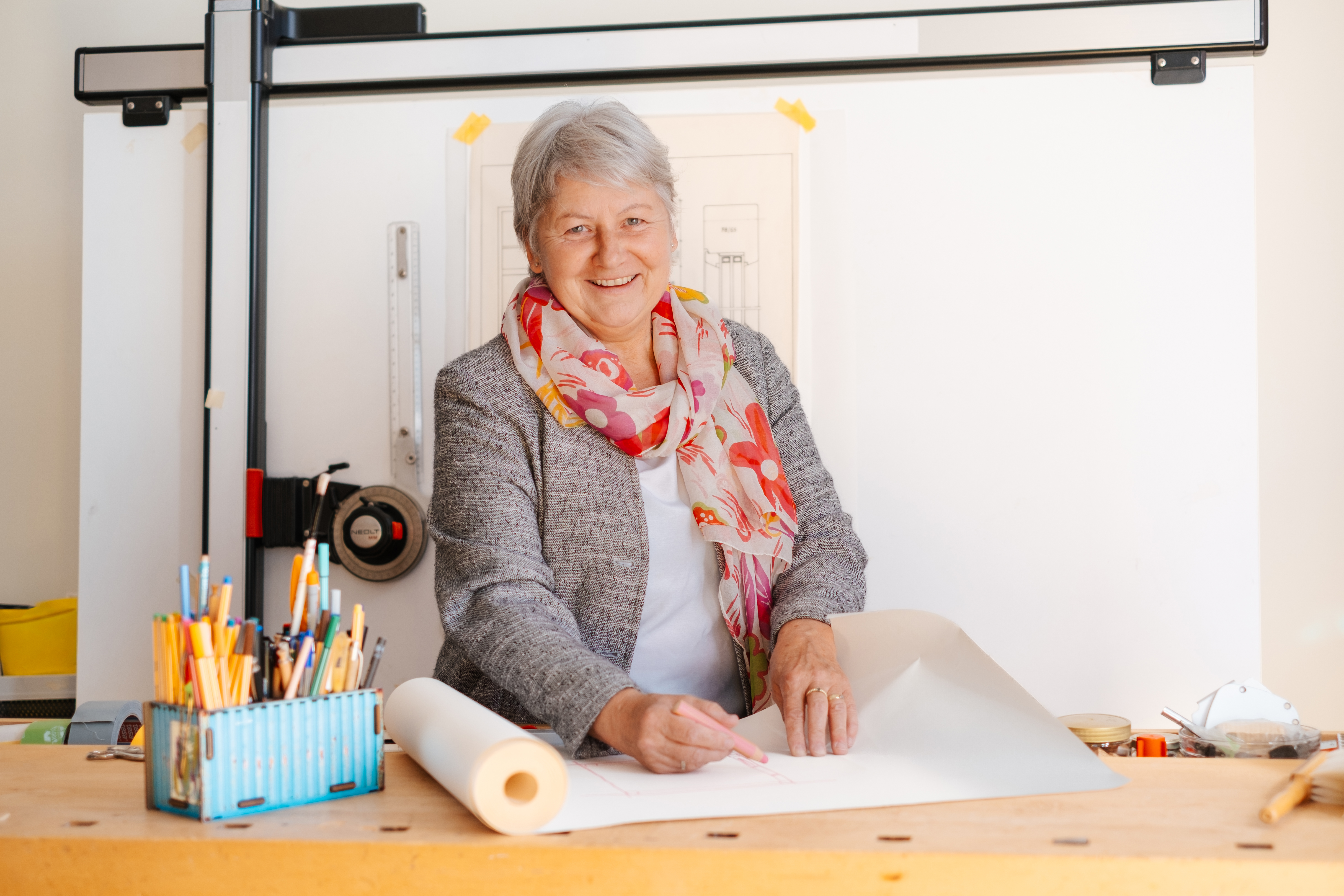Lächelnde Frau mit grauen Haaren steht an einem Zeichentisch und arbeitet mit Bleistift an einer Bauzeichnung; auf dem Tisch liegen Zeichenwerkzeuge und Papierrollen.