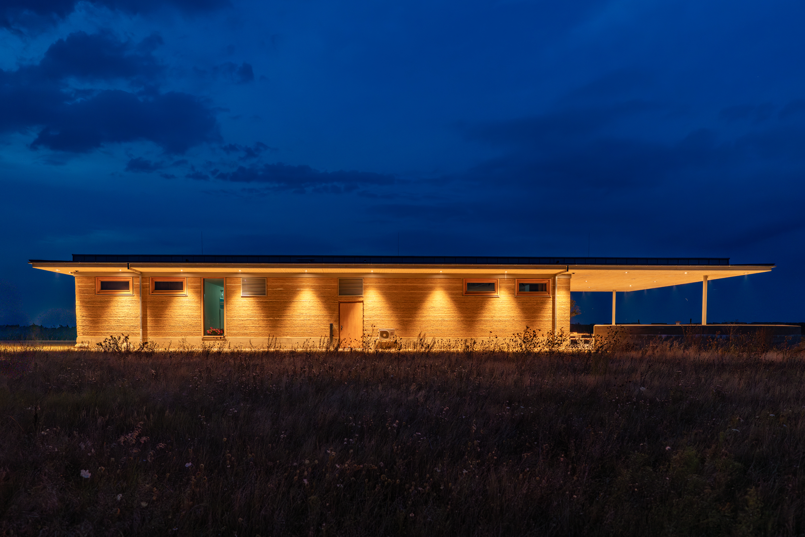 Lange Seitenansicht des Stampflehm-Gebäudes, nachts warm angestrahlt vor tiefblauem Himmel.