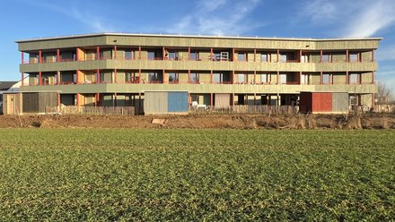 Längliches, dreigeschossiges Wohngebäude mit Holzfassade und durchgehenden Balkonen, freistehend am Rand einer offenen Grünfläche unter blauem Himmel.