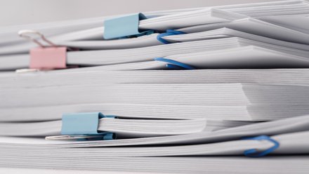 11:47     Nahaufnahme mehrerer Stapel weißer Dokumente auf weißem Hintergrund, mit blauen und rosa Büro- bzw. Foldbackklammern zusammengehalten.