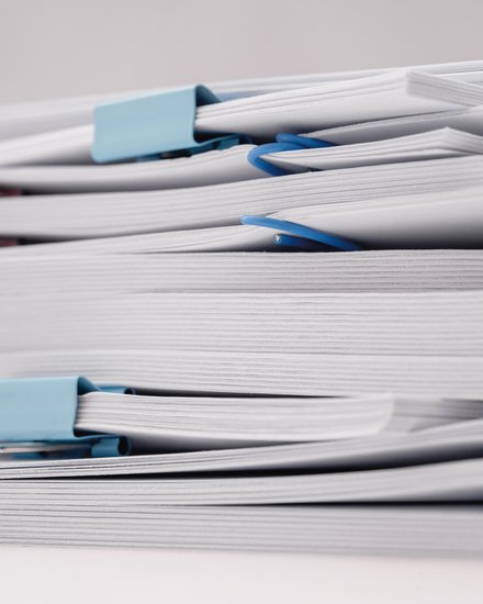 11:47     Nahaufnahme mehrerer Stapel weißer Dokumente auf weißem Hintergrund, mit blauen und rosa Büro- bzw. Foldbackklammern zusammengehalten.
