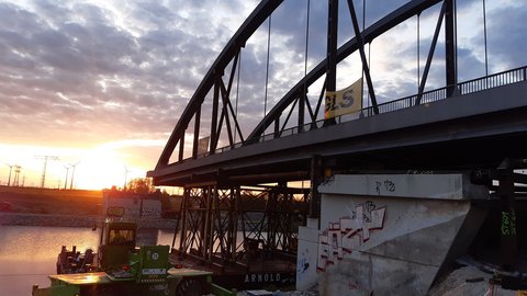 Kuhdammbrücke, Bauphase bei Sonnenuntergang: Stahlbogenbrücke über einem Kanal, Geräte und Ponton unter der Brücke.