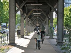 Fahrradfahrer unter einer Stahlbrücke auf einem Radweg mit weißen Markierungen und grünen Bäumen am Rand