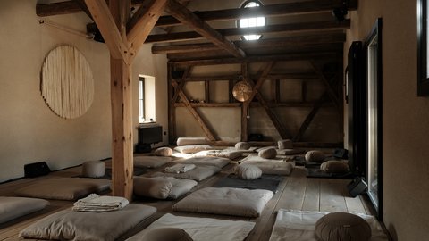  "Weitläufiger Raum mit imposantem Holzfachwerk. Dunkle Holzbalken bilden ein geometrisches Muster. Etwa 15 beige Meditationskissen und Matten sind verteilt auf dem Holzboden angeordnet. An der Wand hängt eine große runde Makramé-Wanddekoration. Ein rundes Fenster mit Gitter oben. Schwarzes Heizgerät sichtbar. Modernes Glasfenster rechts."