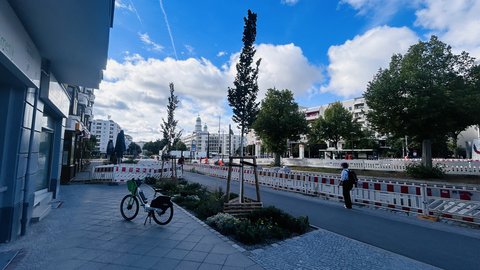 „Blick entlang einer städtischen Straße mit Baustellenabsperrung: Im Vordergrund steht ein Fahrrad neben einem bepflanzten Gehwegstreifen, im Hintergrund ragt ein Denkmal unter blauem Himmel empor.“