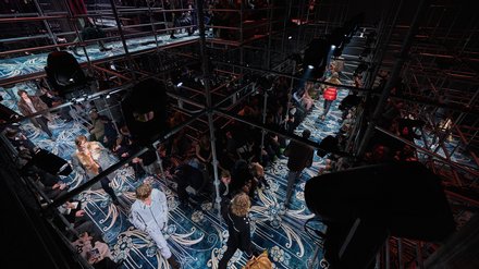 "Ausstellungsraum mit dunkelblauem und cremefarbenem geometrischem Bodenmuster. Die Decke ist schwarz mit industriellen Elementen. Stahlgitterstrukturen und -geländer teilen den Raum in mehrere Ebenen auf. Mehrere schwarze übergroße Kubeskulpturen sind positioniert. Besucher verschiedenen Alters bewegen sich durch die Installation. Projektionen und Videoelemente werden auf Flächen gezeigt. Die Lichtsetzung schafft dramatische Kontraste zwischen Hell und Dunkel."