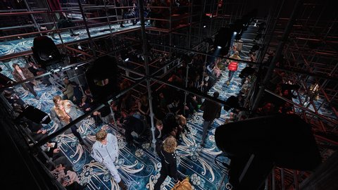 "Ausstellungsraum mit dunkelblauem und cremefarbenem geometrischem Bodenmuster. Die Decke ist schwarz mit industriellen Elementen. Stahlgitterstrukturen und -geländer teilen den Raum in mehrere Ebenen auf. Mehrere schwarze übergroße Kubeskulpturen sind positioniert. Besucher verschiedenen Alters bewegen sich durch die Installation. Projektionen und Videoelemente werden auf Flächen gezeigt. Die Lichtsetzung schafft dramatische Kontraste zwischen Hell und Dunkel."