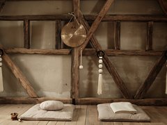 Foto eines rustikalen Yoga- oder Meditationsraums. Sonnenlicht fällt durch ein verborgenes Fenster auf der linken Seite. Decken und Kissen liegen gefaltet auf dem Boden, und ein Gong mit chinesischen Schriftzeichen hängt an einem Holzbalken in der Mitte. Makramee-Seile hängen auf beiden Seiten.