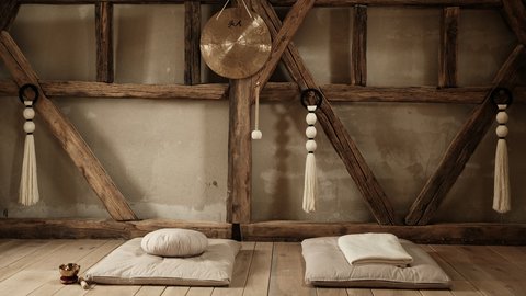 Foto eines rustikalen Yoga- oder Meditationsraums. Sonnenlicht fällt durch ein verborgenes Fenster auf der linken Seite. Decken und Kissen liegen gefaltet auf dem Boden, und ein Gong mit chinesischen Schriftzeichen hängt an einem Holzbalken in der Mitte. Makramee-Seile hängen auf beiden Seiten.