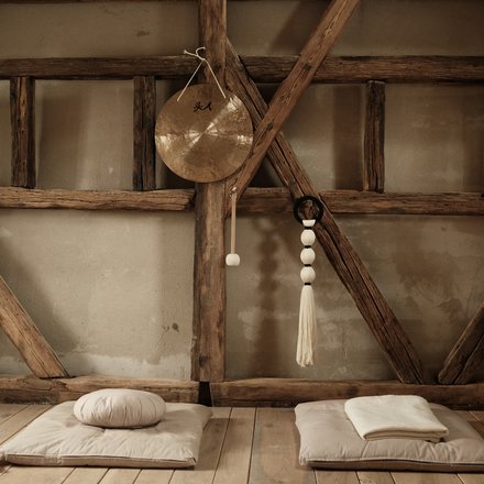 Foto eines rustikalen Yoga- oder Meditationsraums. Sonnenlicht fällt durch ein verborgenes Fenster auf der linken Seite. Decken und Kissen liegen gefaltet auf dem Boden, und ein Gong mit chinesischen Schriftzeichen hängt an einem Holzbalken in der Mitte. Makramee-Seile hängen auf beiden Seiten.