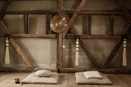 Foto eines rustikalen Yoga- oder Meditationsraums. Sonnenlicht fällt durch ein verborgenes Fenster auf der linken Seite. Decken und Kissen liegen gefaltet auf dem Boden, und ein Gong mit chinesischen Schriftzeichen hängt an einem Holzbalken in der Mitte. Makramee-Seile hängen auf beiden Seiten.