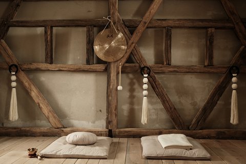 Foto eines rustikalen Yoga- oder Meditationsraums. Sonnenlicht fällt durch ein verborgenes Fenster auf der linken Seite. Decken und Kissen liegen gefaltet auf dem Boden, und ein Gong mit chinesischen Schriftzeichen hängt an einem Holzbalken in der Mitte. Makramee-Seile hängen auf beiden Seiten.