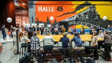 Diskussionsrunde: Menschen sitzen an langem Tisch und diskutieren in einer Veranstaltungshalle.