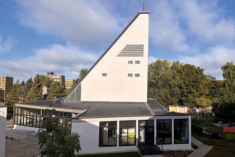 Kirchengebäude mit markantem, schräg ansteigendem Turm und großen Fenstern, umgeben von Bäumen und Wohngebäuden im Hintergrund