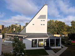Kirchengebäude mit markantem, schräg ansteigendem Turm und großen Fenstern, umgeben von Bäumen und Wohngebäuden im Hintergrund