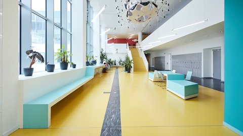 Heller, moderner Schulflur mit großen Fenstern, Sitzbänken, Pflanzen und einer Treppe im Hintergrund