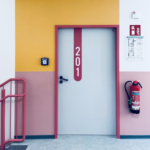 "Heller Flur mit zweifarbiger Wandgestaltung: oben weiß, unten dunkelrosa. Obere Wandhälfte in Gelb. An der Wand links ein blauer Meldekasten. Eine Aufzugstür in Weiß mit dunkelrotem Rahmen zeigt die Nummer '201' in großen weißen Ziffern. Rechts neben der Tür ein roter Feuerlöscher. Ein rotes Metallgeländer ist links sichtbar."