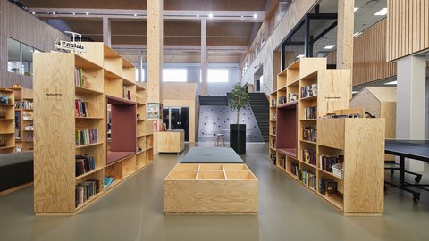 Zentraler Blick durch eine Schulbibliothek mit Bücherregalen aus hellem Holz und Sitznischen. Eine lange Sitzbank steht im Mittelgang; dahinter führen Treppen zu weiteren Ebenen.