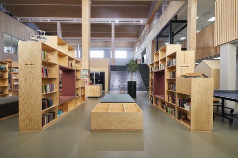 Zentraler Blick durch eine Schulbibliothek mit Bücherregalen aus hellem Holz und Sitznischen. Eine lange Sitzbank steht im Mittelgang; dahinter führen Treppen zu weiteren Ebenen.