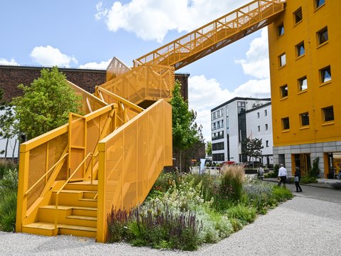 Gelbe Außentreppe mit Brücke in einem begrünten Innenhof zwischen Gebäuden.