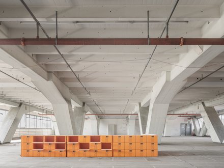 Großer Hallenraum mit orangefarbenem Regalsystem  Ein weiter, leerer Innenraum mit hoher Decke und massiven, weiß gestrichenen Betonstützen. Quer durch den Raum verläuft eine niedrige, lange Struktur aus leuchtend orangefarbenen, würfelförmigen Modulen, die an ein offenes Regalsystem erinnern. Die Halle wirkt roh und industriell, mit sichtbaren Leitungen an der Decke. Das Bild zeigt den baulichen Bestand als offenen, flexiblen Raum. Die orangefarbenen Elemente markieren eine neue Nutzung oder Intervention innerhalb der bestehenden Struktur.