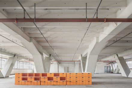 Großer Hallenraum mit orangefarbenem Regalsystem  Ein weiter, leerer Innenraum mit hoher Decke und massiven, weiß gestrichenen Betonstützen. Quer durch den Raum verläuft eine niedrige, lange Struktur aus leuchtend orangefarbenen, würfelförmigen Modulen, die an ein offenes Regalsystem erinnern. Die Halle wirkt roh und industriell, mit sichtbaren Leitungen an der Decke. Das Bild zeigt den baulichen Bestand als offenen, flexiblen Raum. Die orangefarbenen Elemente markieren eine neue Nutzung oder Intervention innerhalb der bestehenden Struktur.
