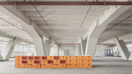 Großer Hallenraum mit orangefarbenem Regalsystem  Ein weiter, leerer Innenraum mit hoher Decke und massiven, weiß gestrichenen Betonstützen. Quer durch den Raum verläuft eine niedrige, lange Struktur aus leuchtend orangefarbenen, würfelförmigen Modulen, die an ein offenes Regalsystem erinnern. Die Halle wirkt roh und industriell, mit sichtbaren Leitungen an der Decke. Das Bild zeigt den baulichen Bestand als offenen, flexiblen Raum. Die orangefarbenen Elemente markieren eine neue Nutzung oder Intervention innerhalb der bestehenden Struktur.
