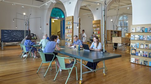 Menschen sitzen an einem langen Tisch in einem hellen Raum mit hohen Bögen und Holzfußboden, im Hintergrund Bücherregale und eine Tafel mit Zeichnung