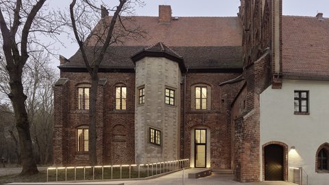 Ein Foto zeigt die Rückseite eines historischen Backsteingebäudes bei Dämmerung. Ein moderner barrierefreier Zugang mit beleuchteten Handläufen führt zu einer Glastür. Der Bau hat ein hohes Ziegeldach, unregelmäßige Fenster, einen runden Vorbau und ein Stuckelement mit einem kleineren Eingang. Hohe, kahle Bäume säumen das Grundstück.