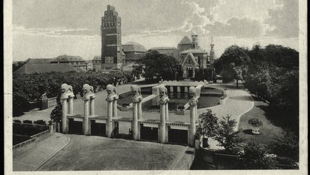 Eine historische Schwarz-Weiß-Fotografie zeigt ein großzügiges Ausstellungsgelände mit einem monumentalen Eingangsportal, mehreren skulpturalen Säulen, einem Wasserbecken und repräsentativen Gebäuden im Hintergrund, darunter ein markanter Turmbau und eine reich verzierte Kapelle, wodurch das Bild die Inszenierung von Architektur, Landschaft und Technik als kulturelles Ereignis dokumentiert und für den historischen Anspruch steht, Baukunst öffentlich zu präsentieren und gesellschaftlich wirksam zu machen.