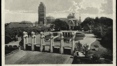 Eine historische Schwarz-Weiß-Fotografie zeigt ein großzügiges Ausstellungsgelände mit einem monumentalen Eingangsportal, mehreren skulpturalen Säulen, einem Wasserbecken und repräsentativen Gebäuden im Hintergrund, darunter ein markanter Turmbau und eine reich verzierte Kapelle, wodurch das Bild die Inszenierung von Architektur, Landschaft und Technik als kulturelles Ereignis dokumentiert und für den historischen Anspruch steht, Baukunst öffentlich zu präsentieren und gesellschaftlich wirksam zu machen.