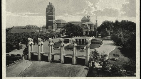 Eine historische Schwarz-Weiß-Fotografie zeigt ein großzügiges Ausstellungsgelände mit einem monumentalen Eingangsportal, mehreren skulpturalen Säulen, einem Wasserbecken und repräsentativen Gebäuden im Hintergrund, darunter ein markanter Turmbau und eine reich verzierte Kapelle, wodurch das Bild die Inszenierung von Architektur, Landschaft und Technik als kulturelles Ereignis dokumentiert und für den historischen Anspruch steht, Baukunst öffentlich zu präsentieren und gesellschaftlich wirksam zu machen.