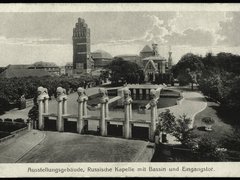 Eine historische Schwarz-Weiß-Fotografie zeigt ein großzügiges Ausstellungsgelände mit einem monumentalen Eingangsportal, mehreren skulpturalen Säulen, einem Wasserbecken und repräsentativen Gebäuden im Hintergrund, darunter ein markanter Turmbau und eine reich verzierte Kapelle, wodurch das Bild die Inszenierung von Architektur, Landschaft und Technik als kulturelles Ereignis dokumentiert und für den historischen Anspruch steht, Baukunst öffentlich zu präsentieren und gesellschaftlich wirksam zu machen.