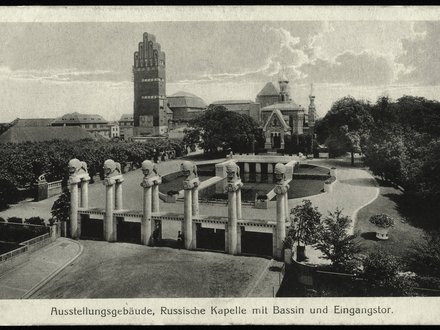 Eine historische Schwarz-Weiß-Fotografie zeigt ein großzügiges Ausstellungsgelände mit einem monumentalen Eingangsportal, mehreren skulpturalen Säulen, einem Wasserbecken und repräsentativen Gebäuden im Hintergrund, darunter ein markanter Turmbau und eine reich verzierte Kapelle, wodurch das Bild die Inszenierung von Architektur, Landschaft und Technik als kulturelles Ereignis dokumentiert und für den historischen Anspruch steht, Baukunst öffentlich zu präsentieren und gesellschaftlich wirksam zu machen.