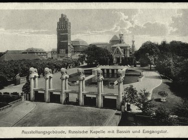 Eine historische Schwarz-Weiß-Fotografie zeigt ein großzügiges Ausstellungsgelände mit einem monumentalen Eingangsportal, mehreren skulpturalen Säulen, einem Wasserbecken und repräsentativen Gebäuden im Hintergrund, darunter ein markanter Turmbau und eine reich verzierte Kapelle, wodurch das Bild die Inszenierung von Architektur, Landschaft und Technik als kulturelles Ereignis dokumentiert und für den historischen Anspruch steht, Baukunst öffentlich zu präsentieren und gesellschaftlich wirksam zu machen.
