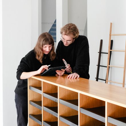 Das Bild zeigt zwei ConcularPersonen in einem hellen Innenraum, die gemeinsam an einem hölzernen Regal oder Sideboard stehen. Beide beugen sich leicht nach vorne und schauen auf ein Tablet, das eine der Personen in der Hand hält, während die andere darauf zeigt. Im Hintergrund sind Leitern und ein neutral gestalteter Raum zu sehen, was an eine Werkstatt, ein Lager oder einen Projektraum erinnert. Die Szene wirkt konzentriert und kollaborativ. Zweck des Bildes ist es, Zusammenarbeit, Planung und praktische Auseinandersetzung mit wiederverwendbaren Bauteilen oder Möbeln im Kontext von ReUse und zirkulärem Bauen darzustellen.