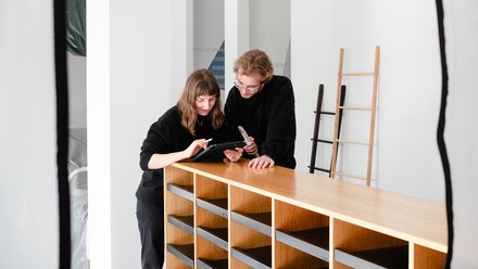 Das Bild zeigt zwei ConcularPersonen in einem hellen Innenraum, die gemeinsam an einem hölzernen Regal oder Sideboard stehen. Beide beugen sich leicht nach vorne und schauen auf ein Tablet, das eine der Personen in der Hand hält, während die andere darauf zeigt. Im Hintergrund sind Leitern und ein neutral gestalteter Raum zu sehen, was an eine Werkstatt, ein Lager oder einen Projektraum erinnert. Die Szene wirkt konzentriert und kollaborativ. Zweck des Bildes ist es, Zusammenarbeit, Planung und praktische Auseinandersetzung mit wiederverwendbaren Bauteilen oder Möbeln im Kontext von ReUse und zirkulärem Bauen darzustellen.