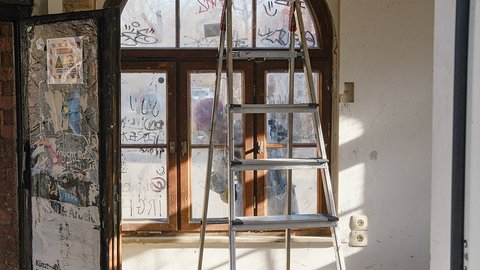 Blick auf ein verwittertes Schaufenster mit Graffiti und Aufklebern. Eine große Leiter steht vor der Fensterfront. Durch die schmuddeligen Fenster ist das Innere eines Raumes sichtbar. Ein Kunstwarnschild ist links an der Wand angebracht."