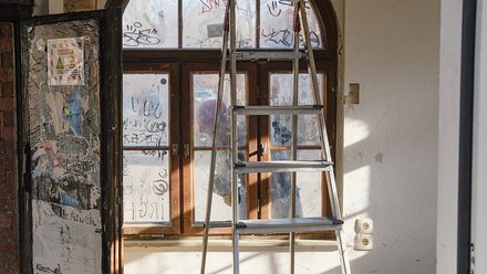 Blick auf ein verwittertes Schaufenster mit Graffiti und Aufklebern. Eine große Leiter steht vor der Fensterfront. Durch die schmuddeligen Fenster ist das Innere eines Raumes sichtbar. Ein Kunstwarnschild ist links an der Wand angebracht."