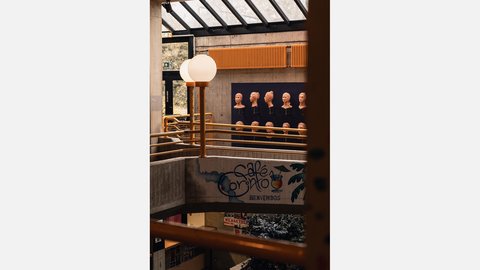 Atrium mit gelben Geländern, runder Leuchte und Betonbrüstung; Wanddisplay mit Tonbüsten; Graffiti „Café Corinto – Bienvenidos“.