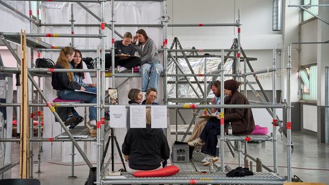 Mehrere Personen sitzen auf einem mehrstöckigen Gerüst in einer Halle und unterhalten sich