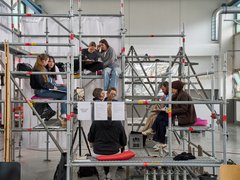 Mehrere Personen sitzen auf einem mehrstöckigen Gerüst in einer Halle und unterhalten sich