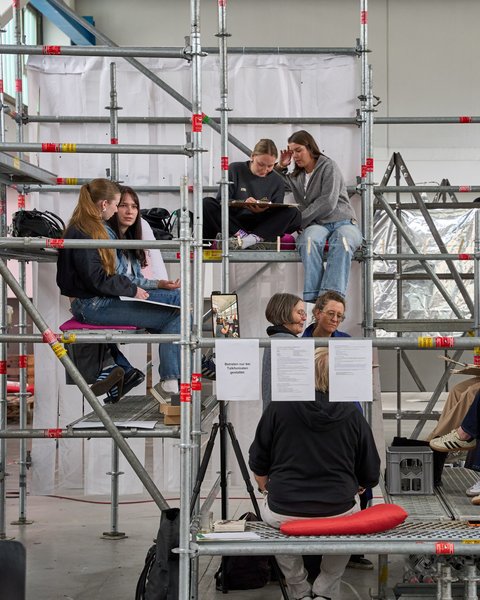 Mehrere Personen sitzen auf einem mehrstöckigen Gerüst in einer Halle und unterhalten sich