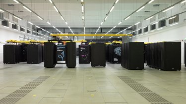 Ein großer, heller Raum mit langen Reihen schwarzer Server-Schränke. Auf einigen Schränken sind Grafiken und Schriftzüge angebracht, etwa zu Hochleistungsrechnen, KI oder Physik. Die Anordnung ist streng symmetrisch. Das Bild zeigt ein modernes Hochleistungsrechenzentrum. Es steht für enorme Rechenkapazitäten, wissenschaftliche Simulationen und datengetriebene Forschung.