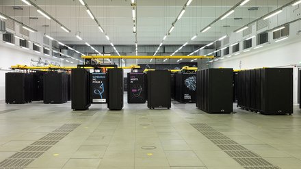 Ein großer, heller Raum mit langen Reihen schwarzer Server-Schränke. Auf einigen Schränken sind Grafiken und Schriftzüge angebracht, etwa zu Hochleistungsrechnen, KI oder Physik. Die Anordnung ist streng symmetrisch. Das Bild zeigt ein modernes Hochleistungsrechenzentrum. Es steht für enorme Rechenkapazitäten, wissenschaftliche Simulationen und datengetriebene Forschung.