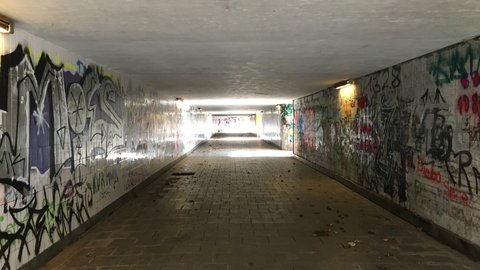 Langer, niedriger Fußgängertunnel mit Graffiti an beiden Wänden und wenigen Deckenleuchten; gepflasterter Boden, Lichtöffnung in der Ferne.
