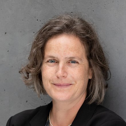 Porträtfoto: Person mit kurzem, leicht gewelltem Haar, lächelt, trägt schwarzen Blazer, gemustertes Oberteil und feine Kette; vor grauer Betonwand, Arme verschränkt.