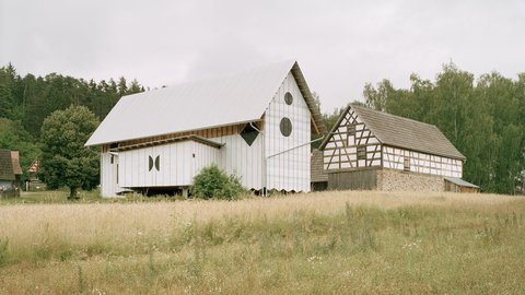 Eine Außenaufnahme, die zwei Gebäude nebeneinander auf einer Wiese zeigt. Links steht das große, neue Holzgebäude, das jetzt mit einer weißen Wetterschutzfolie verkleidet ist und ein glänzendes Metalldach hat. Die markanten runden und rautenförmigen Fensteröffnungen sind mit schwarzer Folie abgedeckt. Rechts daneben steht ein kleineres, historisches Fachwerkgebäude mit einem dunklen Schindeldach. Beide Gebäude stehen vor einem dichten Wald unter einem bewölkten Himmel. Im Vordergrund ist ein Feld mit hohem Gras und Wildblumen.