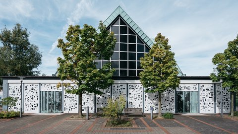 Außenansicht einer modernen Kirche mit markanter Glasgiebelfassade, weiß gestalteter Fassade und Vorplatz mit Bäumen.