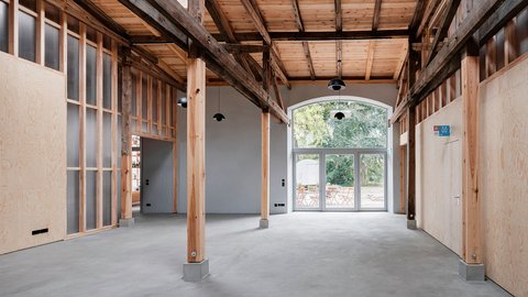 „Großer heller Innenraum mit sichtbarem historischem Holztragwerk, neuen Holzeinbauten und grauem Boden. Eine verglaste Tür führt in den begrünten Außenbereich.“